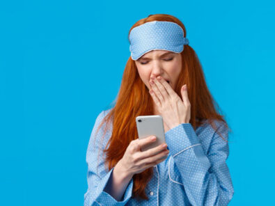 Frau im blauen Schlafanzug mit Augenmaske gähnt mit Smartphone in der Hand